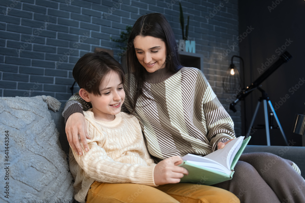 Little boy with his mother sitting on sofa and reading book at home late in evening