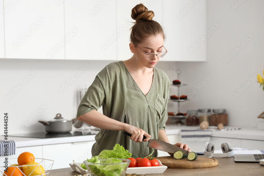 Beautiful young woman cutting cucumber for salad in light kitchen