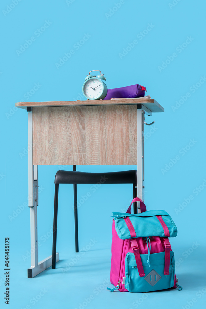 Modern school desk with backpack, alarm clock and stationery on blue background