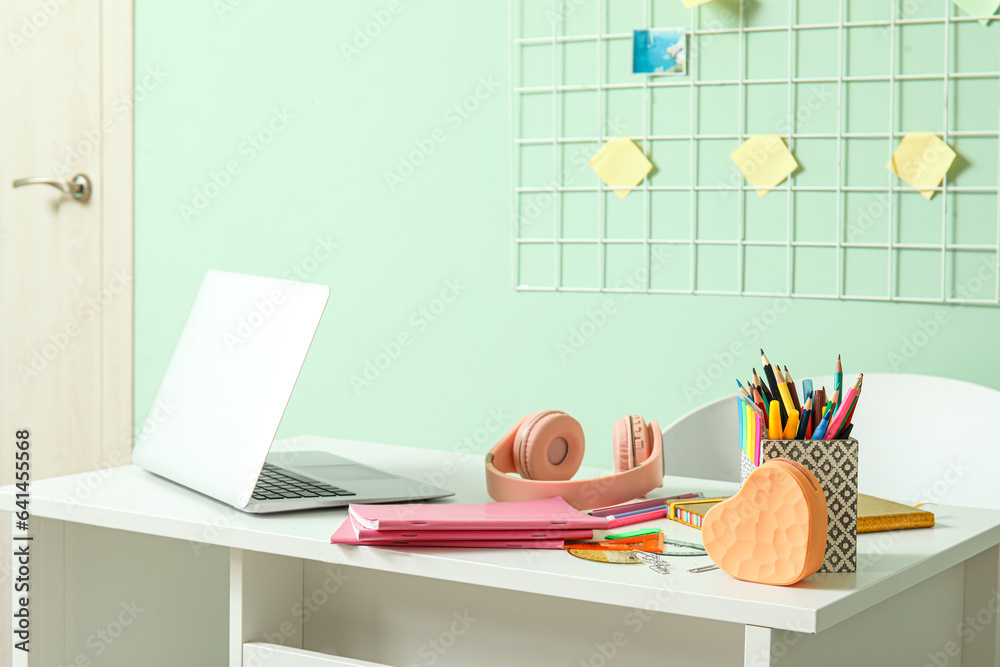 Modern school desk with laptop, headphones and stationery in room near green wall