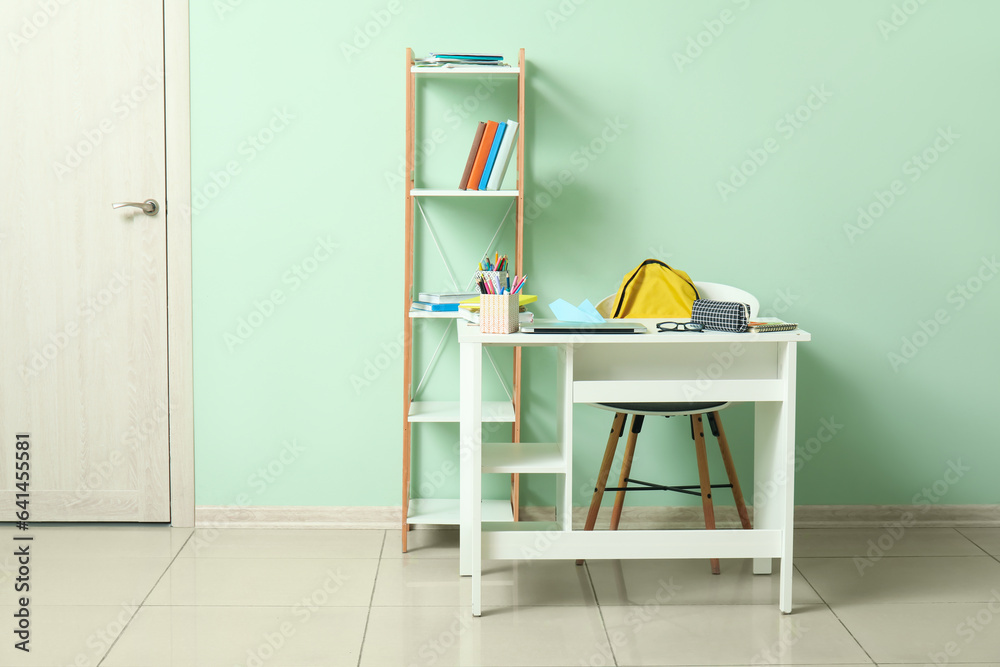 Modern school desk with backpack, laptop and stationery in room near green wall