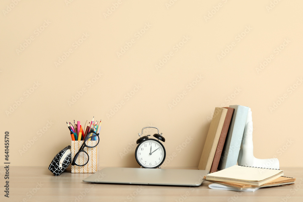 Modern school desk with laptop, alarm clock and stationery in room near beige wall