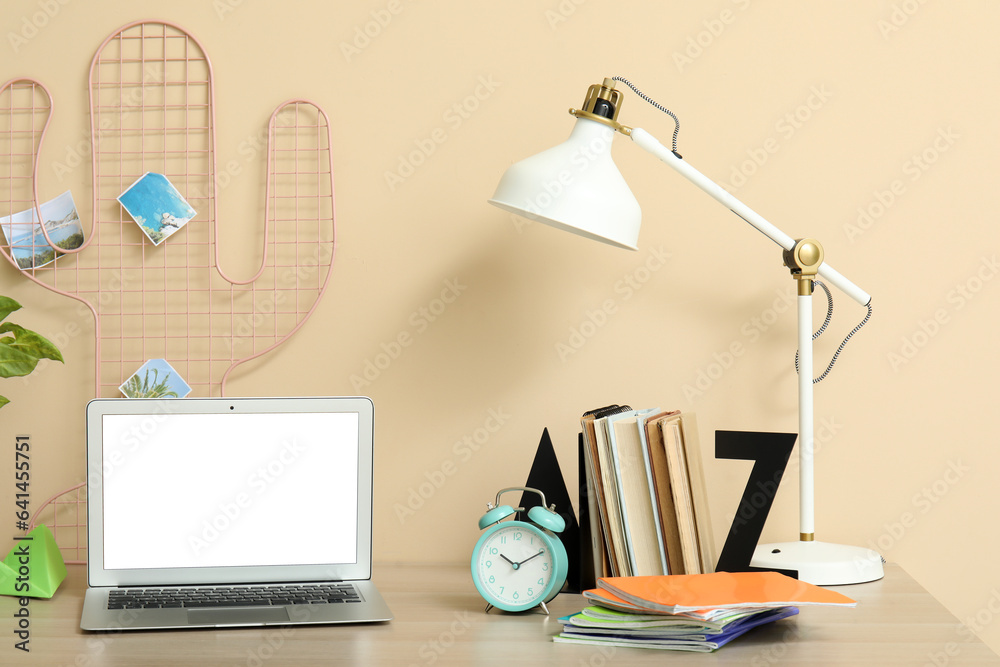 Modern school desk with alarm clock, laptop and stationery in room near beige wall