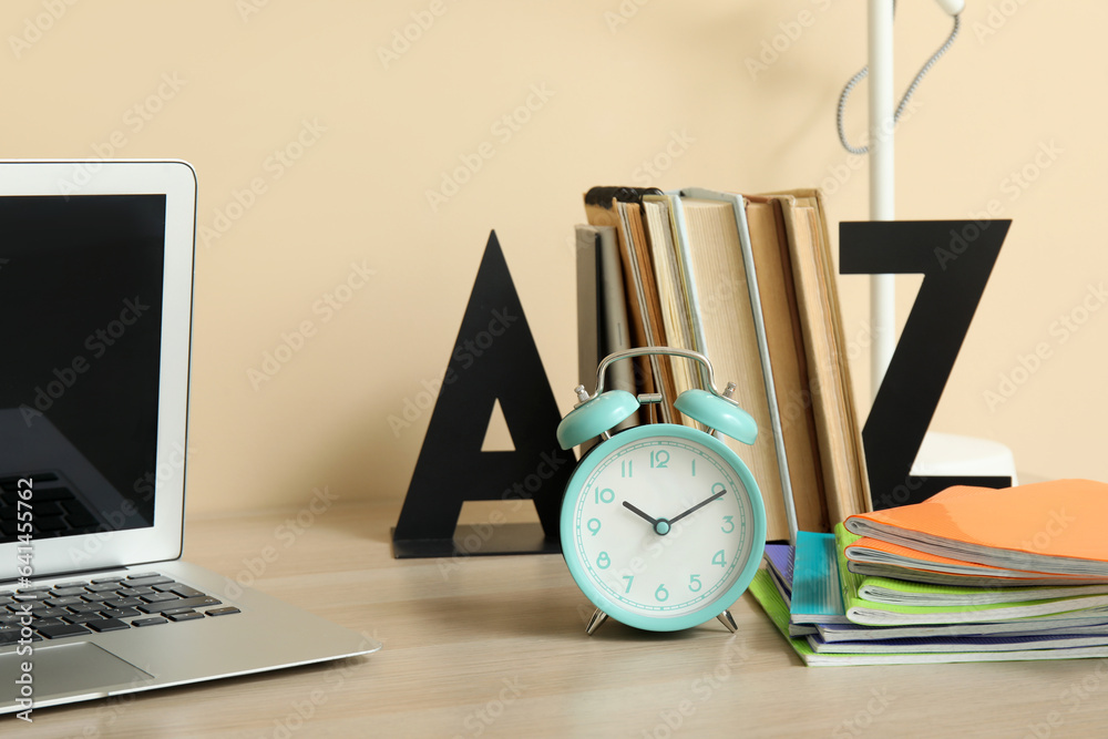 Modern school desk with alarm clock, laptop and stationery in room near beige wall