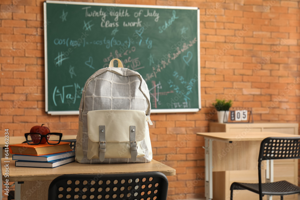 Stylish school backpack with books, apple and eyeglasses on desk in classroom