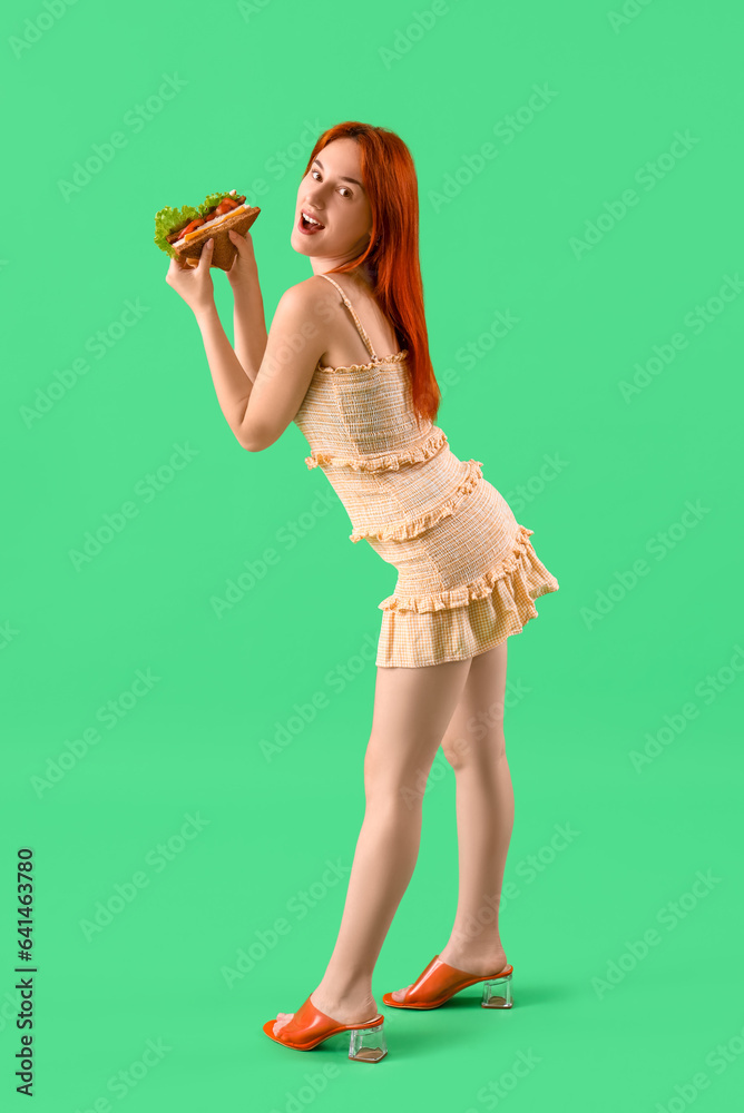 Young redhead woman eating tasty sandwich on green background