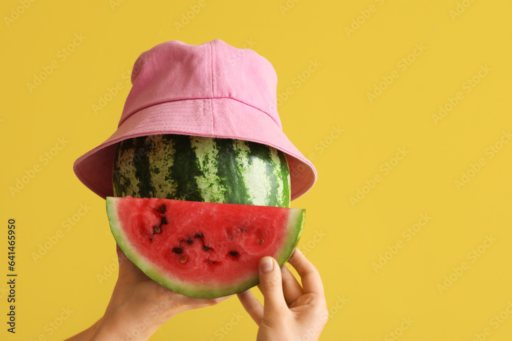 Female hands with whole ripe watermelon with slice and bucket hat on yellow background