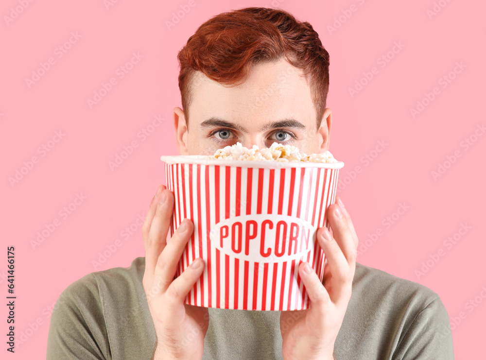 Thoughtful young redhead man with bucket of popcorn on pink background