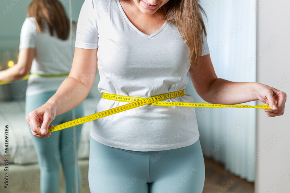 Young woman measuring her waist with tape measure in front of the mirror in her bedroom