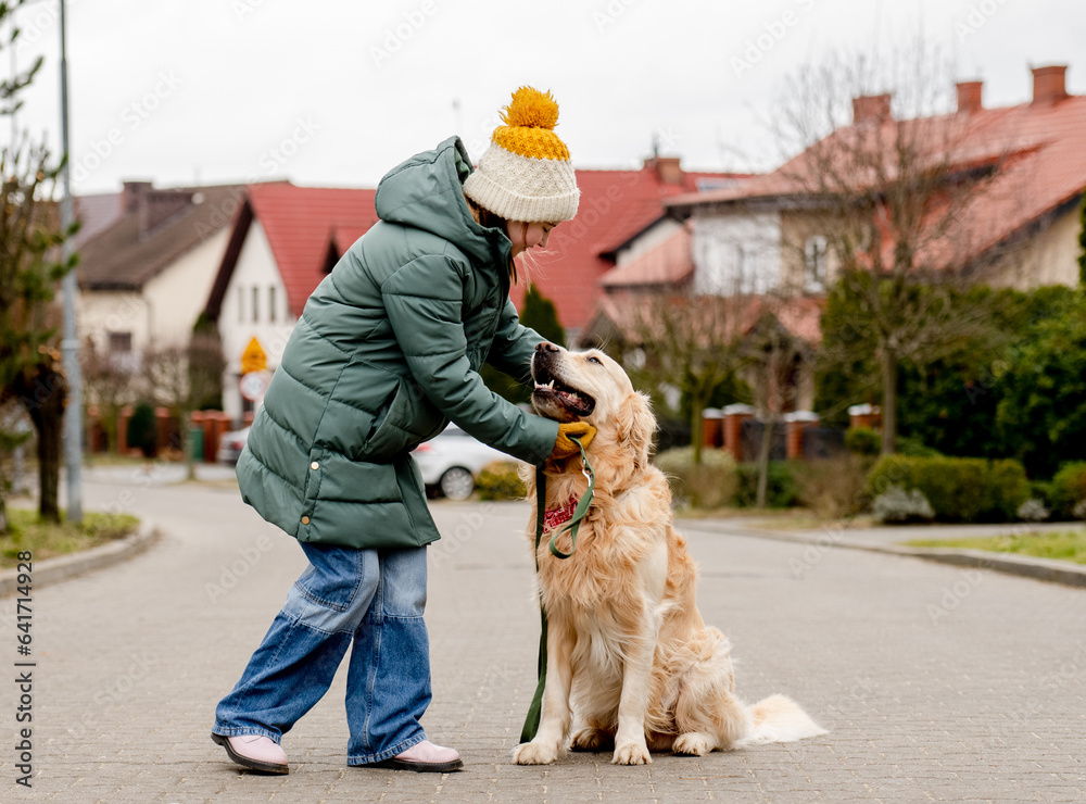 Preteen child girl petting golden retriever dog at autumn street wearing hat and warm jacket. Pretty