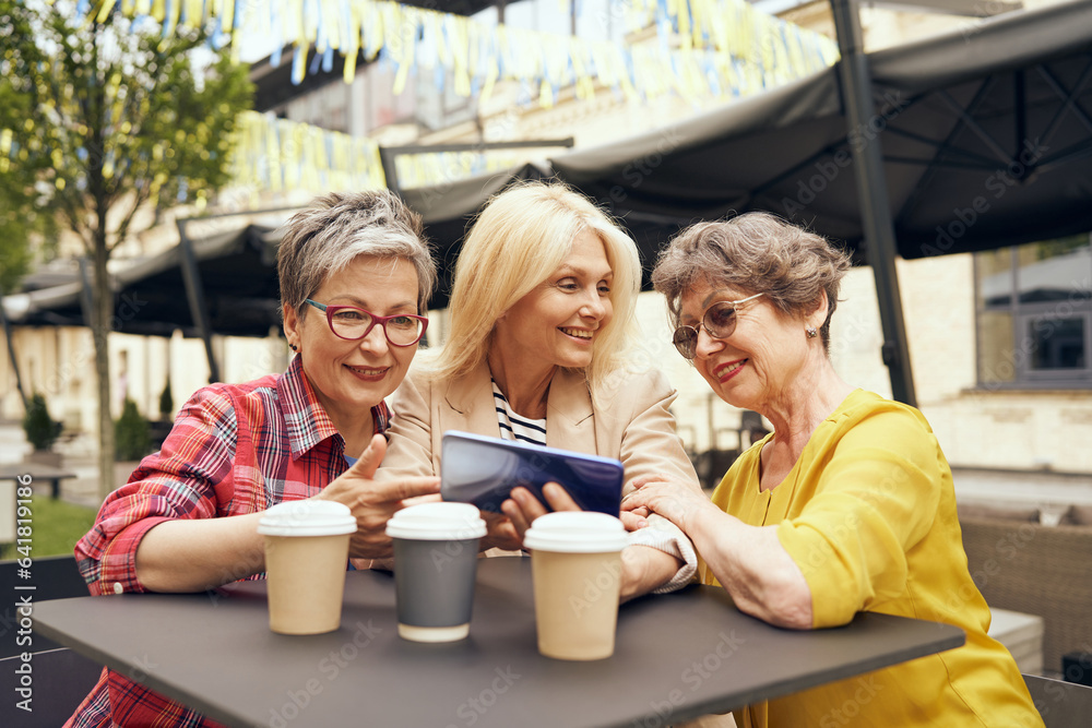 Happy mature women sitting at table with coffee and laughing while watching video on smartphone