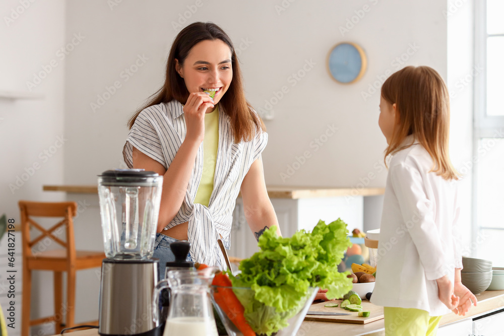 Little girl with her mother eating cucumber in kitchen