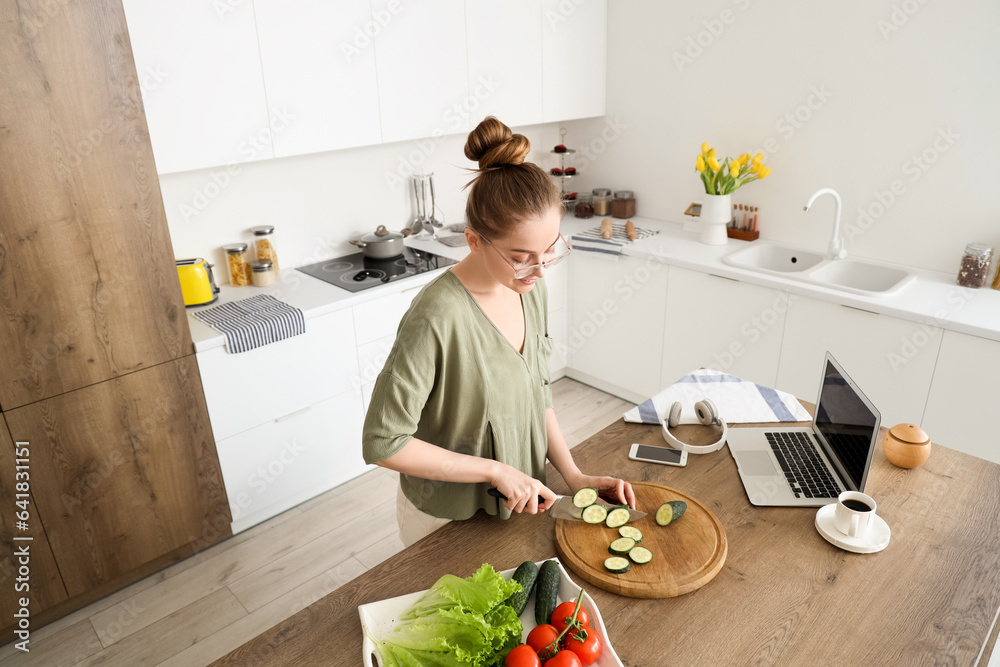 Beautiful young woman cutting cucumber at table with modern laptop in light kitchen