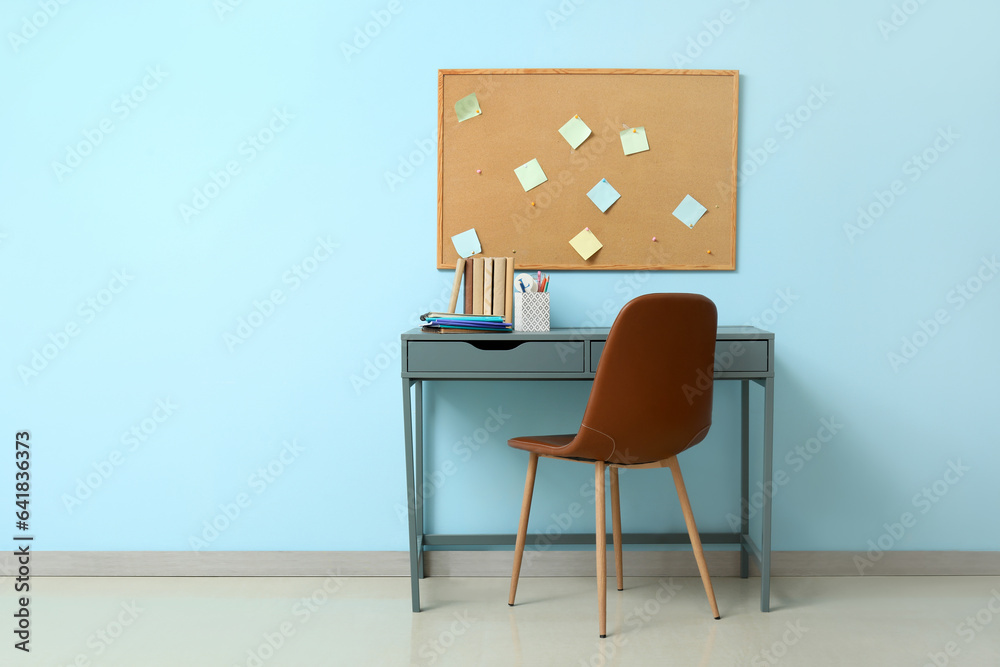 Modern school desk with stationery and cork board in room near blue wall