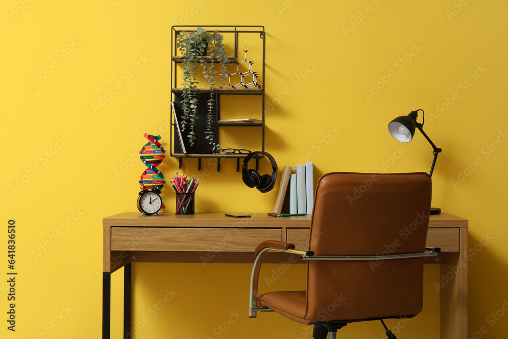 Modern school desk with molecular model, alarm clock and stationery in room near yellow wall