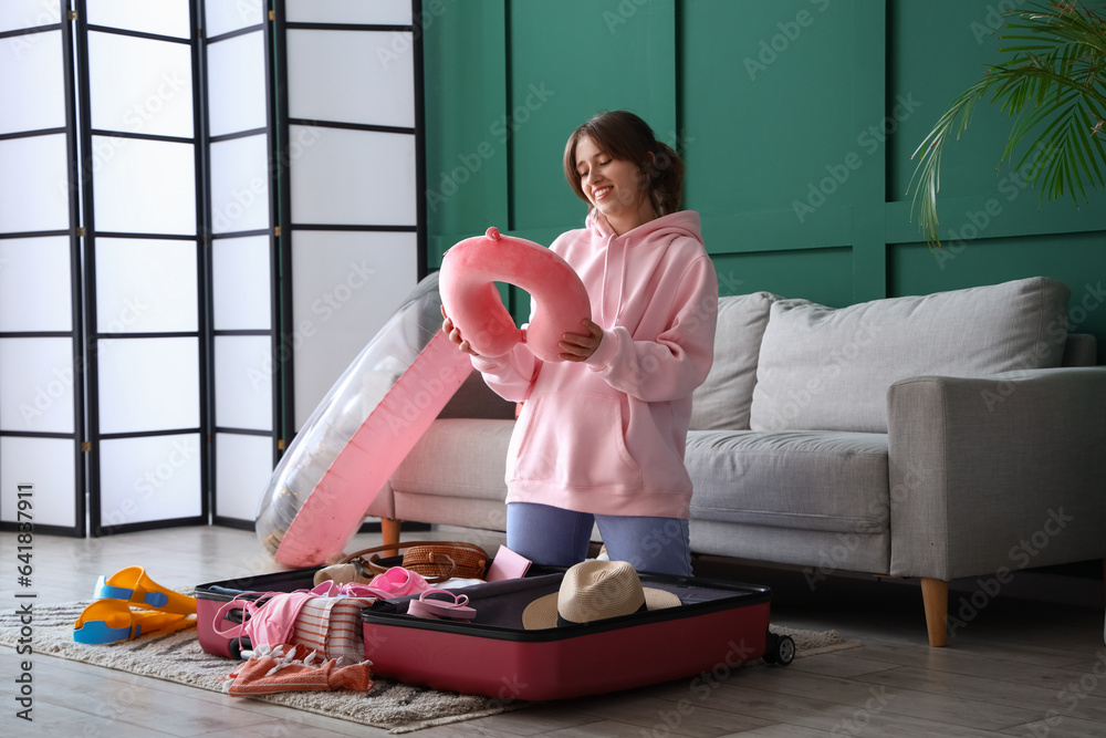 Young woman with neck pillow unpacking suitcase at home