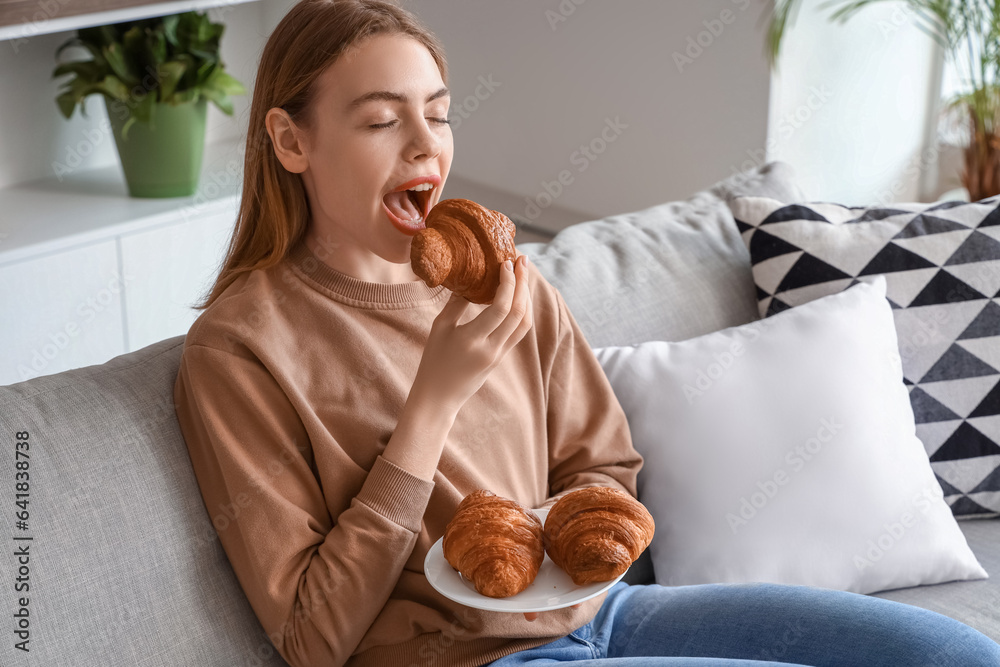 Beautiful young woman eating tasty croissant and sitting on sofa in living room