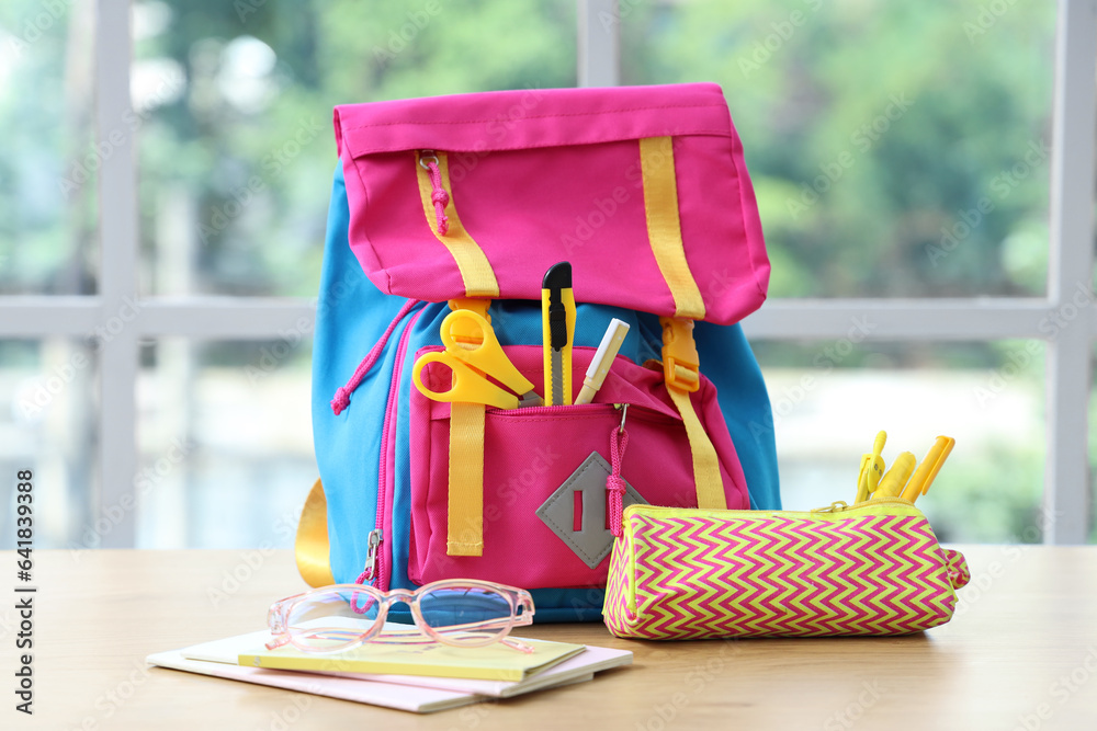 Colorful school backpack with stationery and eyeglasses on wooden table near window in classroom