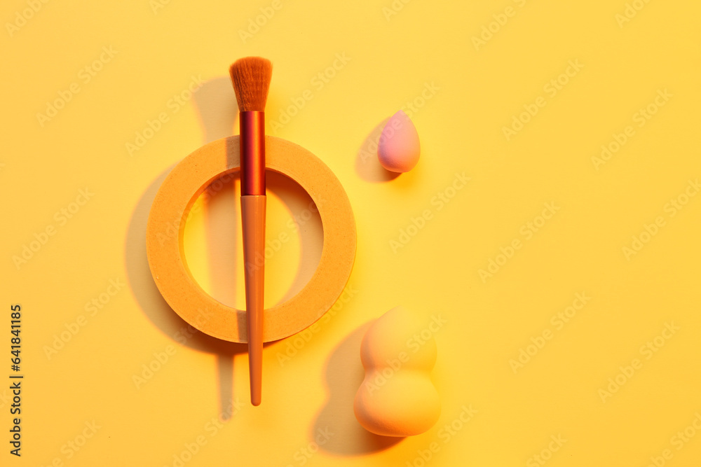 Decorative podium with makeup brush and sponges on yellow background