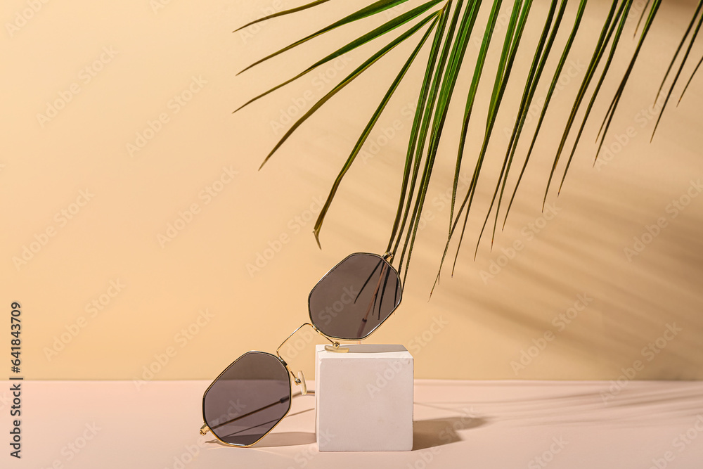 Decorative podium with stylish sunglasses and palm leaf on beige background