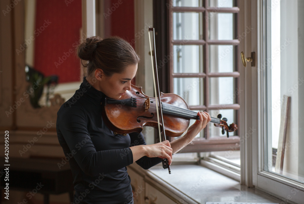 Beautiful woman plays the violin by the window in a classical mansion interior