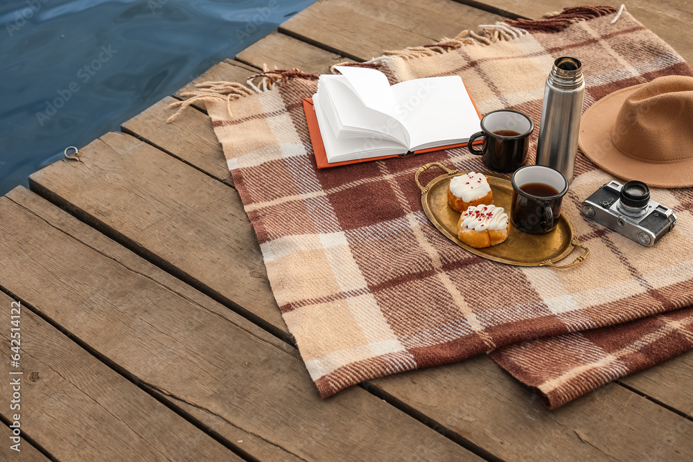 Plaid with book, cups of coffee, thermos, buns, photo camera and hat on bridge near river