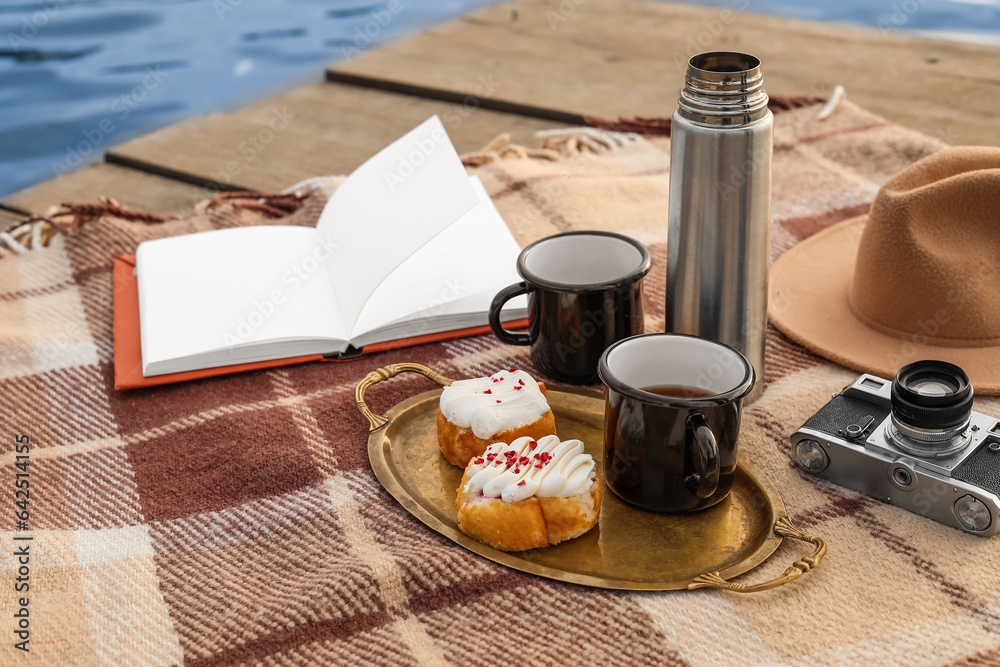 Plaid with book, cups of coffee, thermos, buns, photo camera and hat on bridge near river