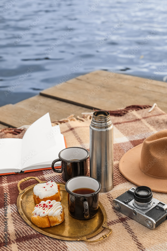 Plaid with book, cups of coffee, thermos, buns, photo camera and hat on bridge near river