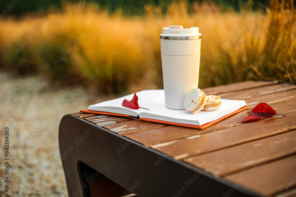 Book with cup of coffee, macaroons and autumn leaves on bench in park at sunset