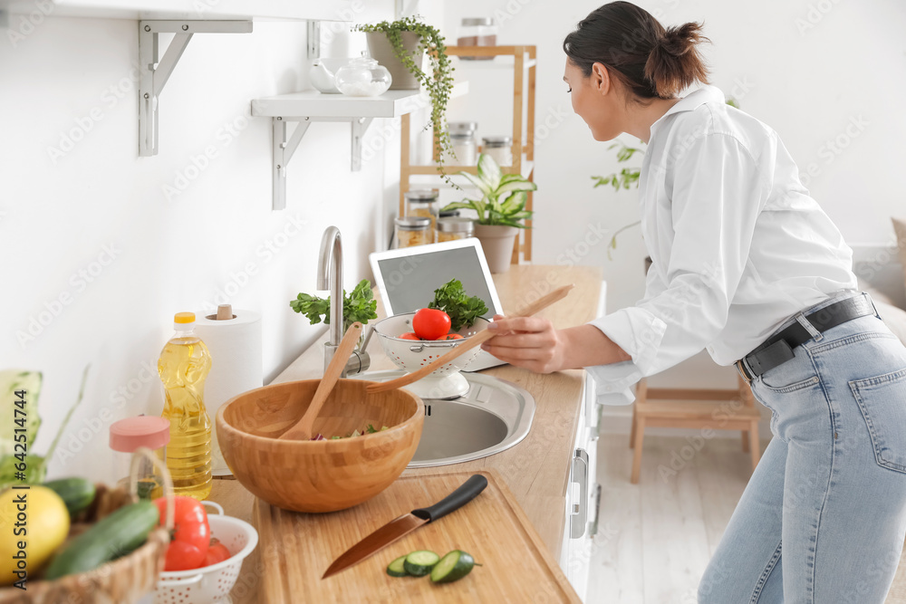 Young woman with laptop making vegetable salad in kitchen