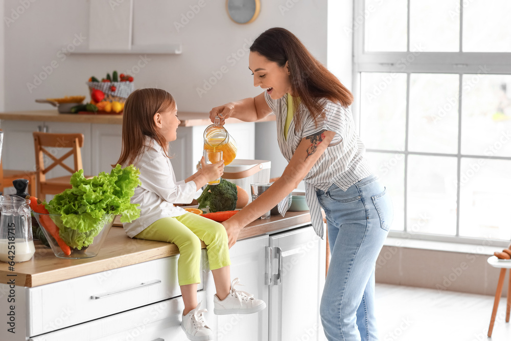 Young woman pouring orange juice into glass of her little daughter in kitchen