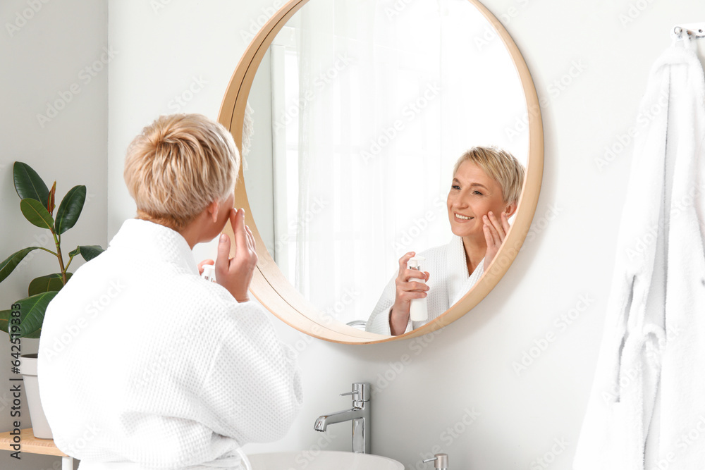 Mature woman applying facial cream near mirror in bathroom