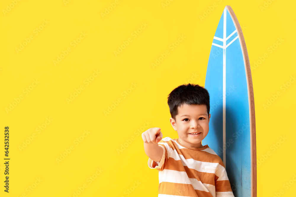 Cute little Asian boy with surfboard pointing at camera on yellow background