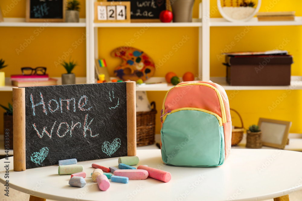 School backpack, chalkboard with text HOMEWORK and crayons on table in room