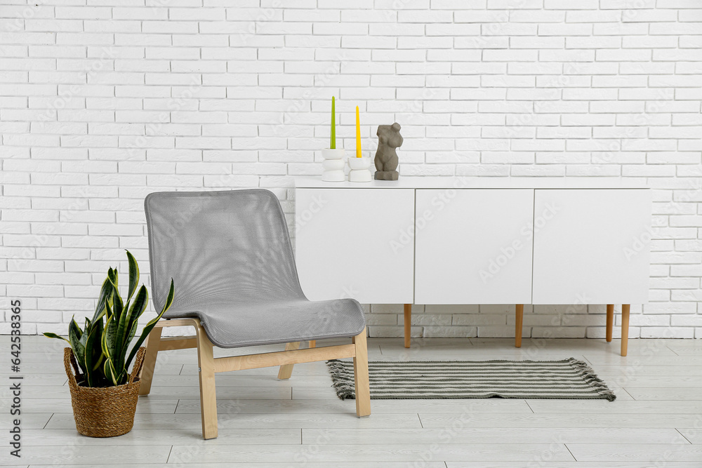 Stylish armchair, houseplant and cabinet with decor near light brick wall