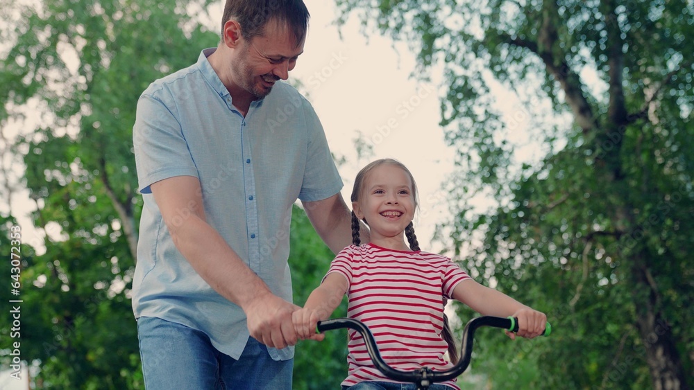 Happy family, little girl in helmet together with her dad learns to ride bicycle outdoors in summer.