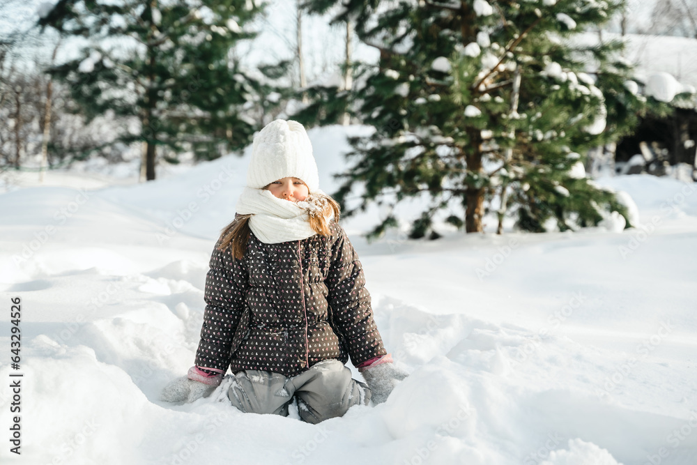 Little cute girl in white hat,scarf snood is lying,playing in snow. Kid walking in beautiful frozen 