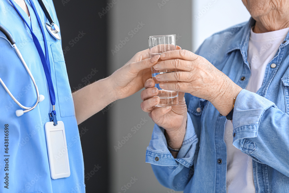 Female caregiver giving glass of water to senior woman in room, closeup