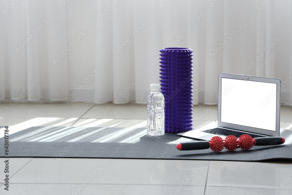 Laptop with bottle of water and rollers on fitness mat in gym