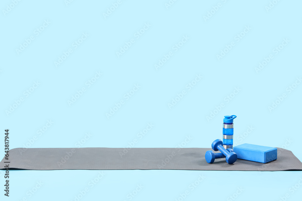 Fitness mat with bottle of water, block and dumbbells on blue background