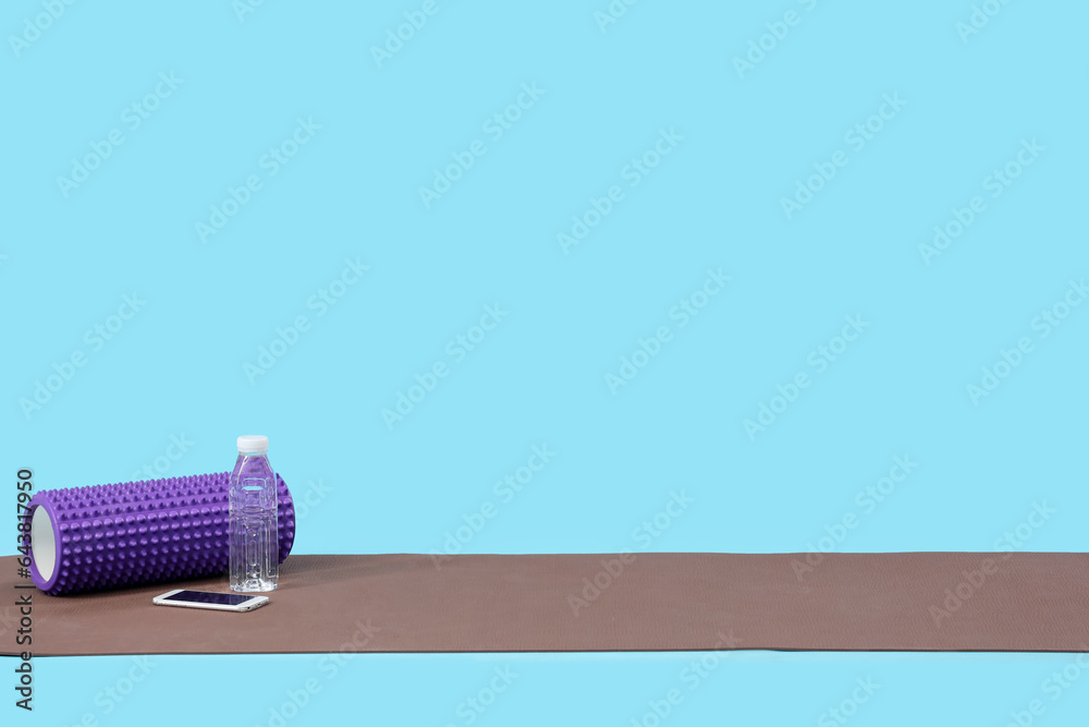 Fitness mat with foam roller, mobile phone and bottle of water on blue background