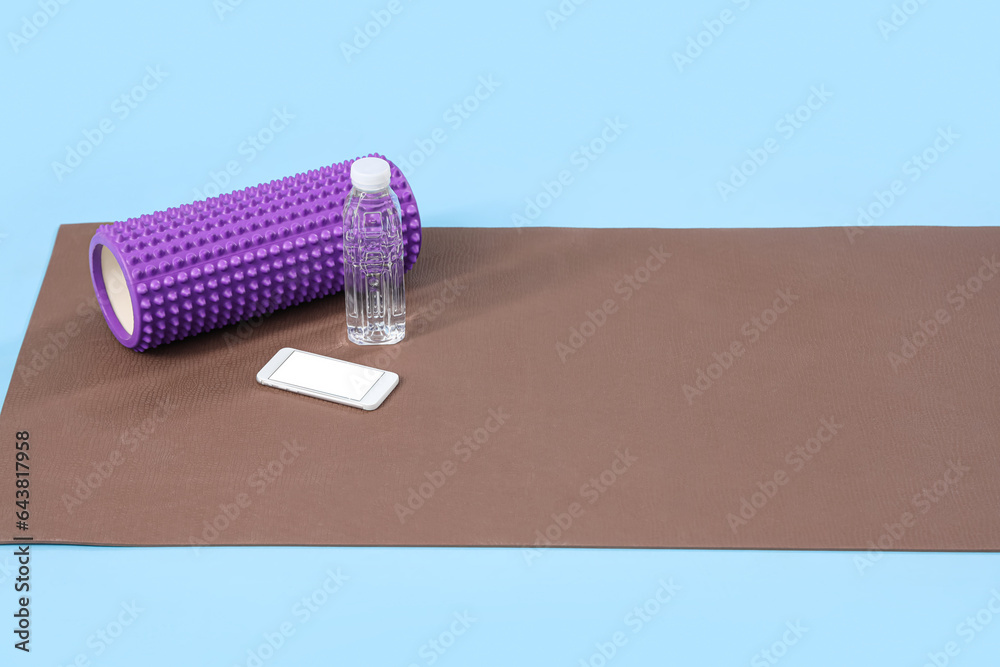 Fitness mat with foam roller, mobile phone and bottle of water on blue background