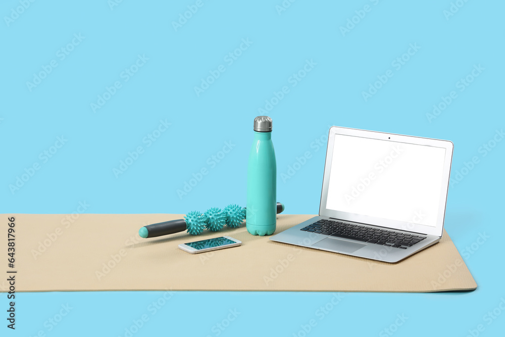 Fitness mat with laptop, mobile phone, bottle and massage roller on blue background