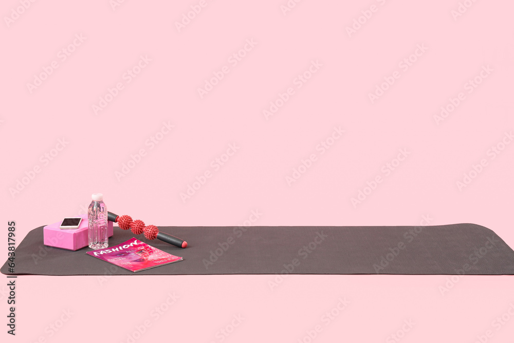 Fitness mat with sports equipment, mobile phone, magazine and bottle of water on pink background