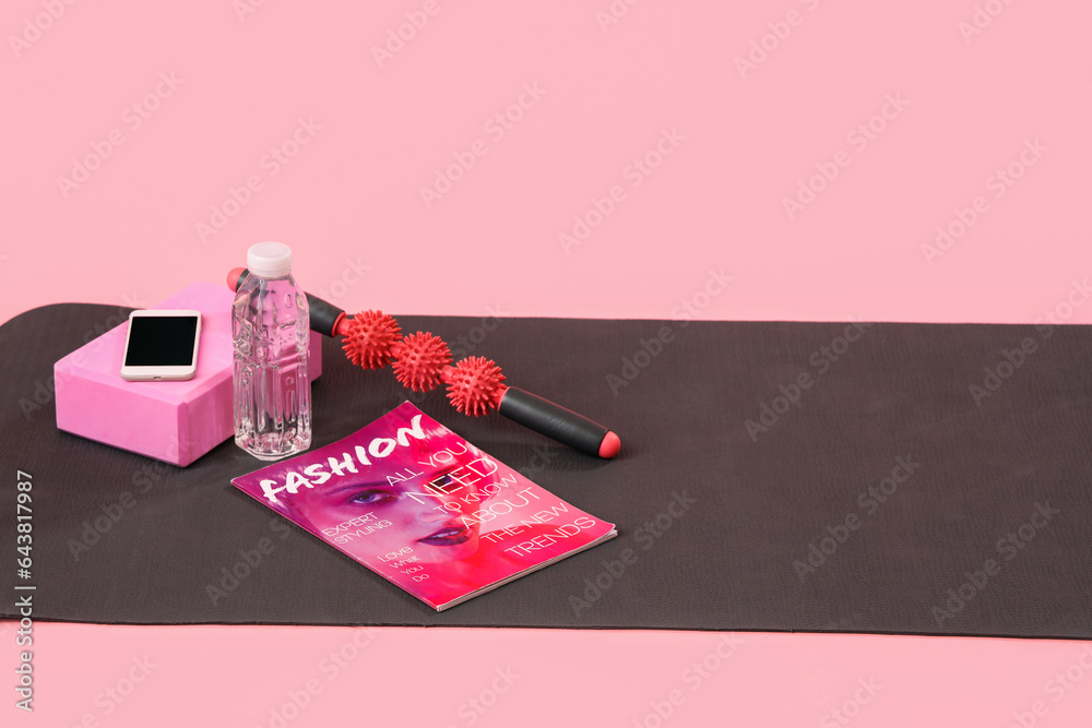 Fitness mat with sports equipment, mobile phone, magazine and bottle of water on pink background