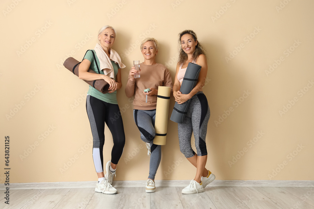 Sporty mature women with yoga mats near beige wall