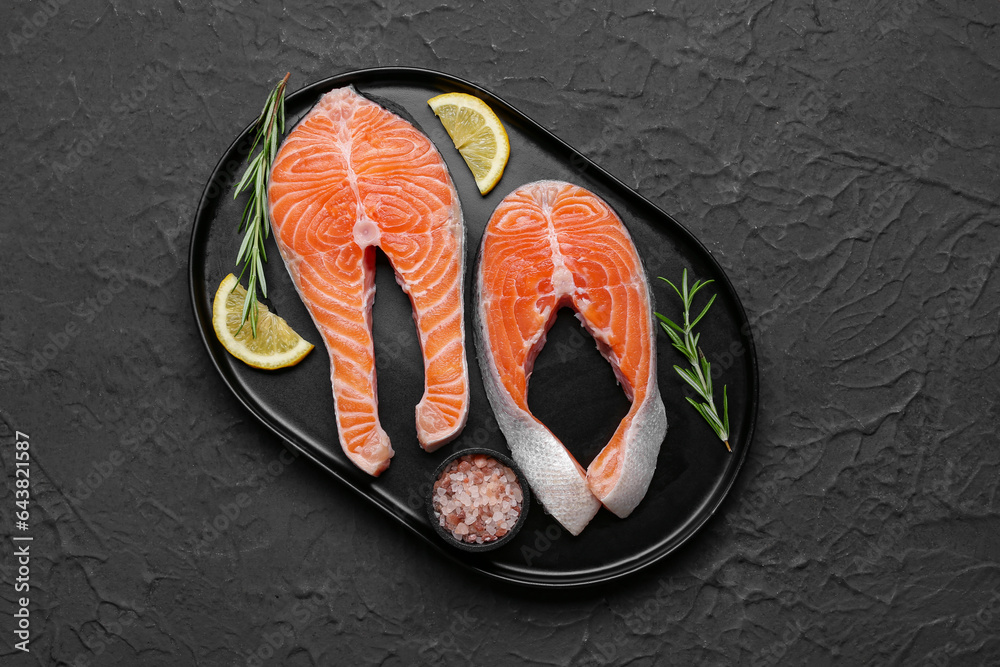 Plate with raw salmon steaks and different spices on black background