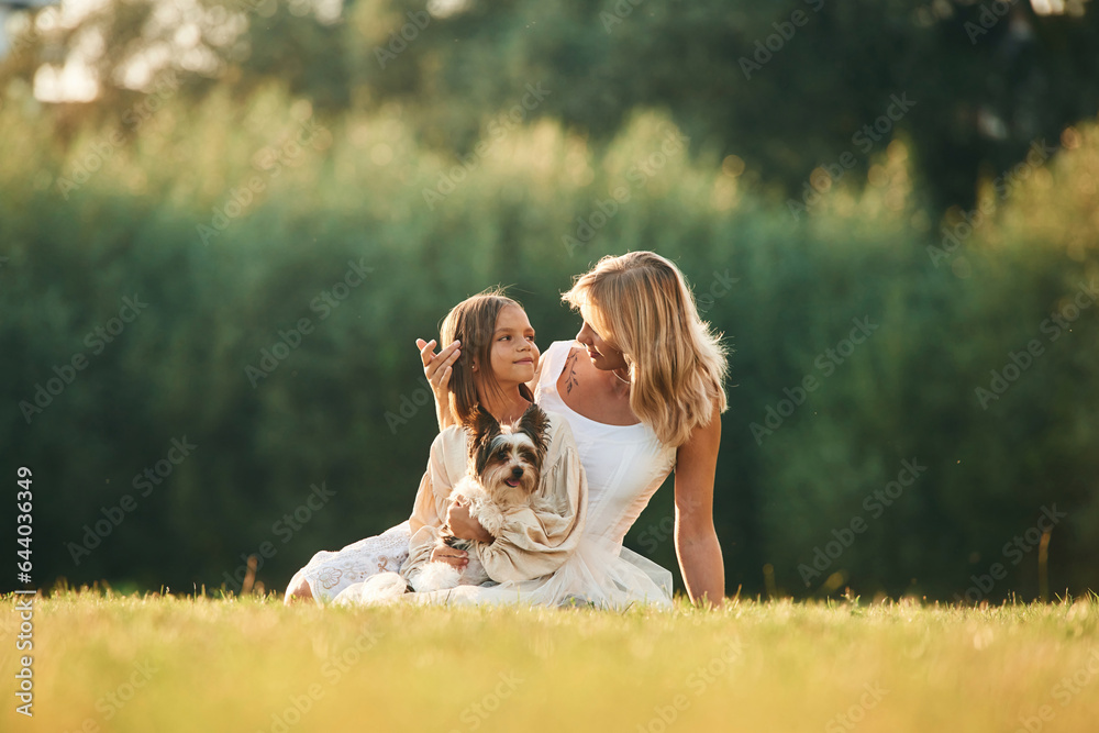 Sitting on the ground. Mother with her daughter and cute dog are on the field outdoors