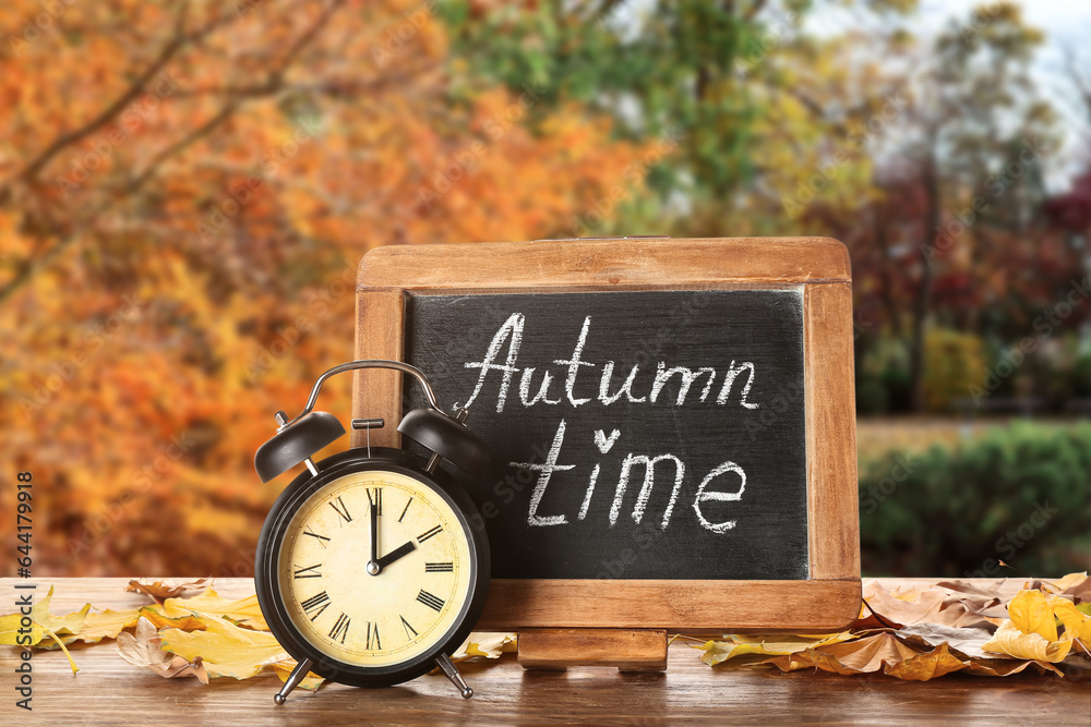 Alarm clock, chalkboard with text AUTUMN TIME and leaves on table outdoors