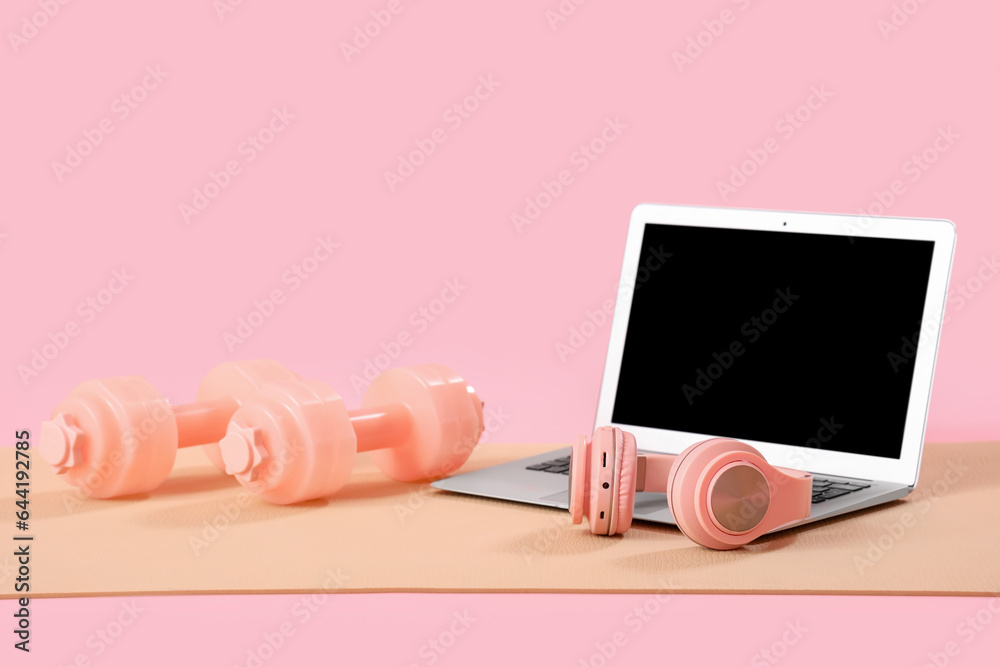 Fitness mat with laptop, headphones and dumbbells on pink background